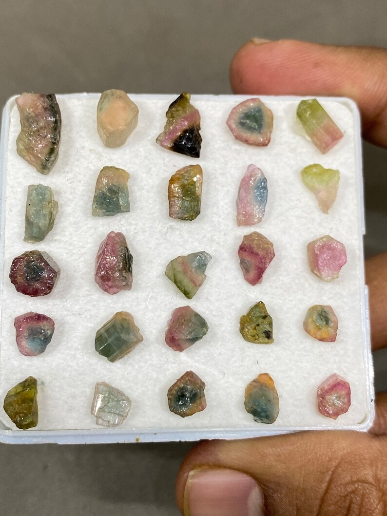 May include: A collection of 24 small, colourful, raw tourmaline crystals in a white foam-lined box. The crystals are various shades of pink, green, and black.