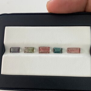 May include: Five rectangular cut gemstones in a black display case. The gemstones are arranged in a row from left to right. The gemstones are various shades of pink, green, and blue.