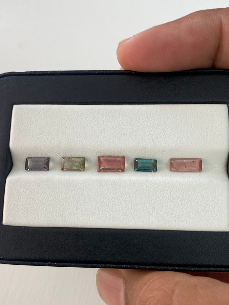 May include: Five rectangular cut gemstones in a black display case. The gemstones are arranged in a row from left to right. The gemstones are various shades of pink, green, and blue.