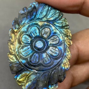 May include: A carved labradorite gemstone with a floral design. The stone features a central flower surrounded by detailed leaves, with iridescent blue, green, and gold hues. The gemstone is held in a hand.