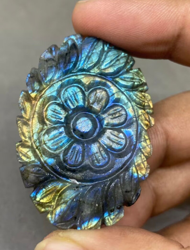 May include: A carved labradorite gemstone with a floral design. The stone features a central flower surrounded by detailed leaves, with iridescent blue, green, and gold hues. The gemstone is held in a hand.