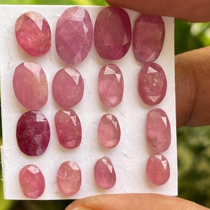 May include: A collection of 15 pink oval-shaped gemstones on a white card. The gemstones are faceted and have a translucent appearance.