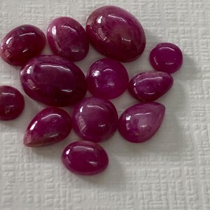 Puede incluir: Una colección de once piedras preciosas cabujón pulidas de color rojo rosado intenso, de diversos tamaños y formas, incluyendo óvalos, redondas y gotas, cada una con una superficie lisa y reflectante que muestra inclusiones naturales.