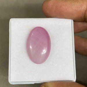May include: A pale pink oval cabochon gemstone with a smooth, polished surface. The stone is displayed in a white box.