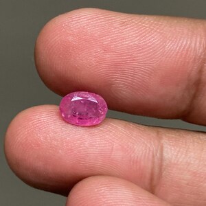 Op de afbeelding: Een kleine, ovaalvormige, roze edelsteen. De edelsteen wordt tussen de vingers gehouden en toont zijn gepolijste oppervlak en levendige kleur. De steen lijkt een robijn te zijn, met een licht doorschijnende kwaliteit.