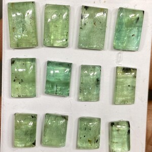 May include: A collection of twelve rectangular, translucent green gemstones. Each gem has a slightly different shade of green, with some showing darker inclusions. The gems are arranged on a white surface, showcasing their clarity and color variations.
