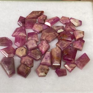 May include: A collection of faceted, raw ruby gemstones in various shapes and sizes. The gems display a range of pink and reddish-brown hues, with some having translucent areas. They are presented in a white tray, showcasing their natural beauty.