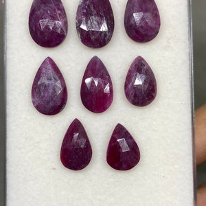Puede incluir: Ocho piedras preciosas de rubí facetadas en forma de pera en una caja de presentación blanca. Los rubíes son de color rojo intenso con un ligero tono púrpura.