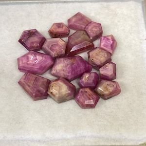 May include: A collection of faceted, hexagonal gemstones in shades of pink and red. The gems have a polished, translucent appearance, with visible internal patterns. They are arranged on a white surface within a small tray.