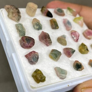 May include: A collection of 20+ small, colourful, and translucent gemstones in a white box. The gemstones are various shades of pink, green, and brown.