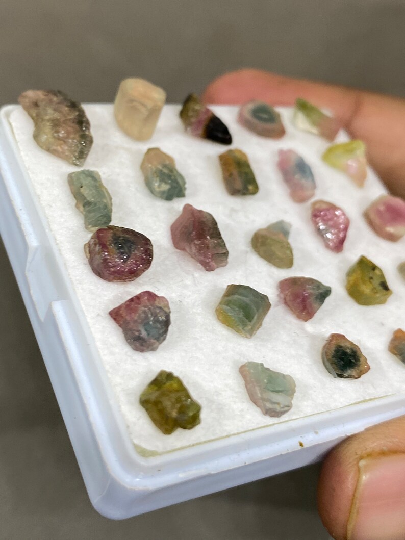 May include: A collection of 20+ small, colourful, and translucent gemstones in a white box. The gemstones are various shades of pink, green, and brown.