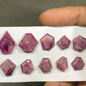 May include: A collection of ten faceted, pinkish-red gemstones arranged in two rows on a white surface. Each stone features a distinct geometric shape with visible internal banding and varying shades of deep magenta and lighter pink hues.