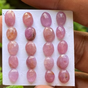 May include: A collection of 18 pink faceted oval-shaped gemstones on a white card. The gemstones are arranged in three rows of six.