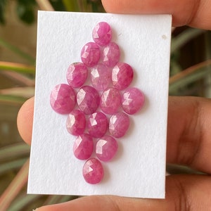 May include: A collection of 15 pink ruby gemstones, each with a faceted, rounded shape. The stones are arranged in a diamond pattern on a white card.