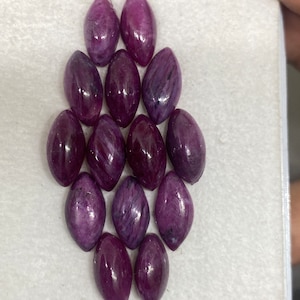 Puede incluir: Una colección de 12 cabujones de piedras preciosas púrpuras en forma de marquesa. Las piedras preciosas están dispuestas en forma de diamante sobre una superficie blanca.