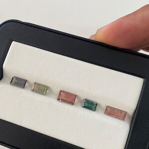 May include: Six rectangular gemstones in a white display case. The gemstones are arranged in a row from left to right. The gemstones are various shades of blue, green, pink, and purple.