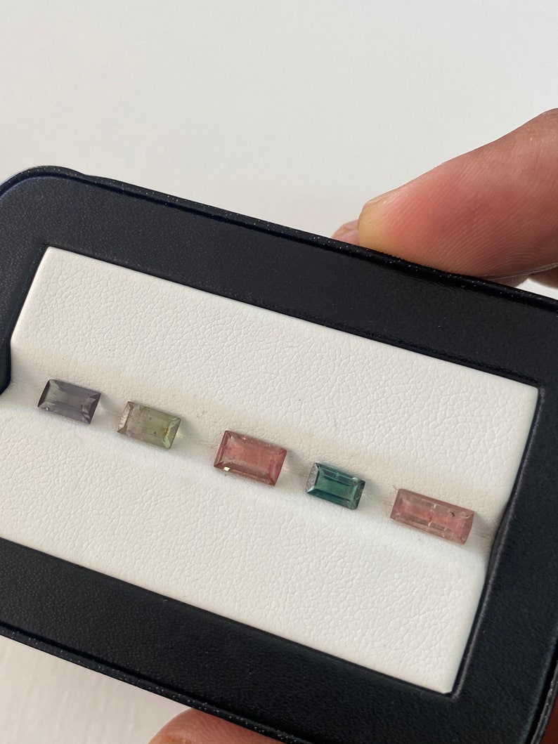 May include: Six rectangular gemstones in a white display case. The gemstones are arranged in a row from left to right. The gemstones are various shades of blue, green, pink, and purple.
