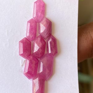 May include: A collection of faceted, pink gemstones arranged on a white surface. The gemstones have a hexagonal shape with multiple facets, reflecting light. The color is a vibrant pink, with variations in tone across the stones. The arrangement suggests a jewelry component.