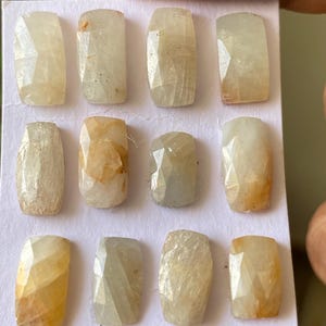 Pode incluir: Uma coleção de doze pedras preciosas retangulares facetadas, em tons variáveis de amarelo pálido e creme. As gemas estão dispostas em um cartão branco, mostrando seus cortes únicos e variações de cor natural. As pedras são seguradas pelos dedos de uma pessoa.