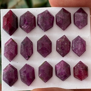 May include: Twelve faceted purple gemstones arranged in a grid on a white surface. The gemstones are all the same size and shape, and they have a slightly iridescent sheen.