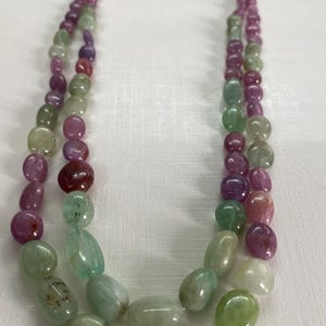 May include: A two-strand necklace featuring a variety of colorful, polished gemstones. The beads are in shades of pink, purple, green, and clear. The necklace is strung on a gold-colored cord.