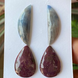 Könnte beinhalten: Vier facettierte Edelsteine. Zwei haben eine Halbmondform mit einem Farbverlauf von Blau nach Grau. Die anderen beiden sind tropfenförmig, tief burgunderfarben und mit sichtbaren Einschlüssen. Alle werden auf einer weißen Oberfläche präsentiert.