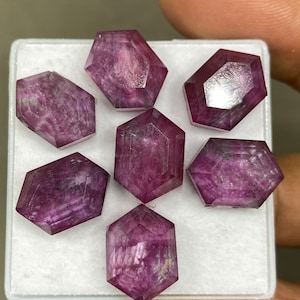 Puede incluir: Seis piedras preciosas facetadas de color rosa púrpura en una caja blanca. Las piedras preciosas están talladas en octógonos con un acabado pulido.