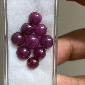 May include: A collection of nine polished, cabochon-cut rubies. The gemstones are a deep, rich red color and are displayed in a clear plastic case with a white fabric lining. The rubies are round and have a smooth, glossy surface.