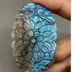 May include: An oval-shaped, carved gemstone with a floral design. The stone features a central flower surrounded by detailed petals and leaves. The color transitions from a light, translucent hue to vibrant blues and greens.