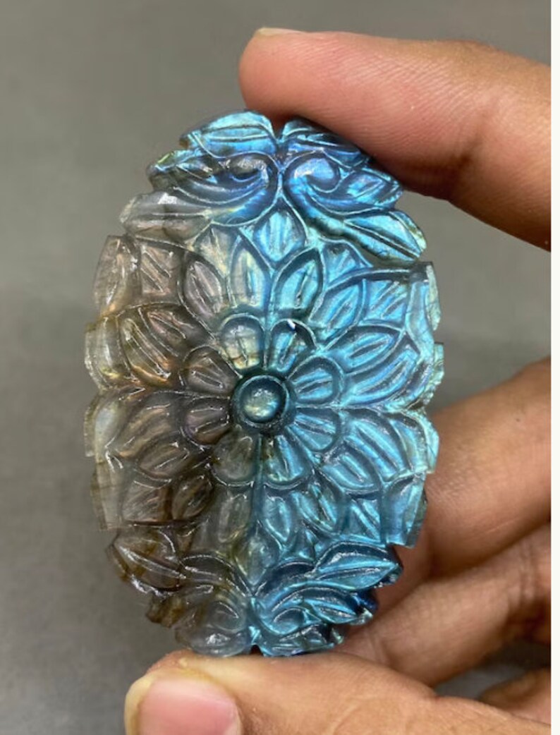 May include: An oval-shaped, carved gemstone with a floral design. The stone features a central flower surrounded by detailed petals and leaves. The color transitions from a light, translucent hue to vibrant blues and greens.