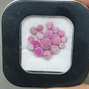 May include: A selection of small, round, pink gemstones presented in a black square container with a white interior. The gemstones range in shades of pink, from pale to deep magenta.