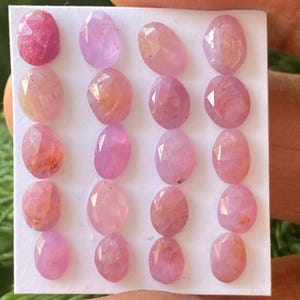 Puede incluir: Una colección de 18 gemas facetadas de color rosa y morado dispuestas en una tarjeta blanca. Las gemas son de forma ovalada y tienen una variedad de tonos de rosa y morado.