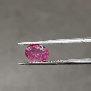 Op de afbeelding: Een kleine, ovaalvormige, roze edelsteen wordt vastgehouden door een zilveren pincet. De edelsteen heeft een enigszins onregelmatig oppervlak en lijkt doorschijnend. De achtergrond is effen, neutraal grijs.