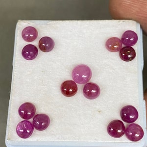 Op de afbeelding: Een verzameling ronde, gepolijste edelstenen in verschillende tinten roze en rood. De edelstenen worden gepresenteerd op een wit oppervlak, vastgehouden door een hand. De edelstenen, mogelijk robijnen, zijn cabochons van verschillende grootte.