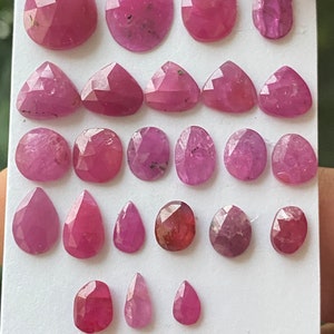Puede incluir: Una colección de 25 piedras preciosas de rubí rosa de varias formas y tamaños, incluyendo cortes de pera, ovalados y triangulares. Las piedras preciosas se muestran sobre un fondo blanco.