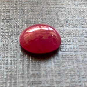 Op de afbeelding: Een gepolijste, ovale, robijnrode edelsteen. De cabochon-geslepen steen heeft een glad, afgerond oppervlak dat licht reflecteert, waardoor een glanzend uiterlijk ontstaat. De edelsteen ligt op een getextureerd, grijs oppervlak.