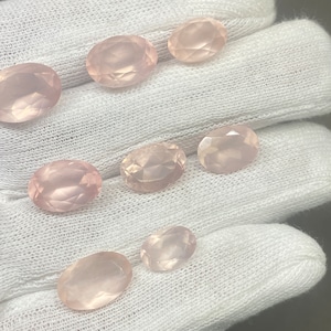 Könnte beinhalten: Eine Sammlung oval geschliffener Rosenquarz-Edelsteine. Die Edelsteine haben eine zarte rosa Farbe und eine facettierte Oberfläche. Die Steine werden auf weißem Hintergrund präsentiert.