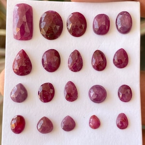 May include: A collection of 18 faceted ruby gemstones in various shapes and sizes, including oval, pear, and rectangle, displayed on a white card.