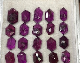 Puede incluir: Una colección de 15 piedras preciosas de color púrpura facetadas en una caja blanca. Las piedras preciosas están todas cortadas en una forma similar, con una base hexagonal y una parte superior puntiaguda.