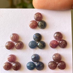 May include: A collection of faceted gemstones in various colors, including pink, blue, and dark green. The gemstones are arranged in small groups on a white surface, showcasing their polished surfaces and unique color variations.