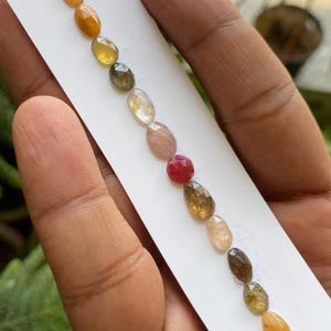 Puede incluir: Una colección de gemas facetadas de forma ovalada en varios colores, incluyendo amarillo, naranja, marrón, rosa y rojo, dispuestas en una tarjeta blanca. Las gemas están pulidas. La imagen está tomada con luz natural, mostrando los colores.