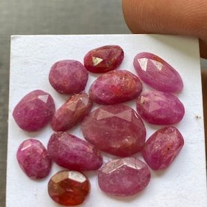 Puede incluir: Una colección de doce piedras preciosas facetadas de forma ovalada en tonos rosa y rosa rojizo, dispuestas sobre una tarjeta cuadrada blanca. Cada piedra presenta una superficie pulida con múltiples facetas que refleja la luz.