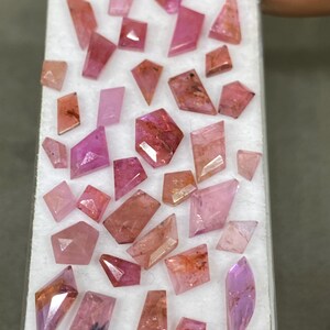 May include: A collection of faceted gemstones in shades of pink, red, and purple. The gemstones are various shapes, including triangles, rectangles, and irregular forms. They are displayed on a white surface, held by a hand.