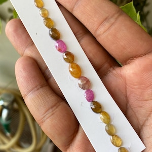 Peut inclure: Une collection de pierres précieuses de forme ovale, dans les tons de jaune, marron et rose, est disposée sur une carte blanche. Les pierres précieuses sont tenues dans une main, mettant en valeur leurs surfaces facettées et leurs couleurs variées.