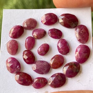 Puede incluir: Una colección de 18 piedras preciosas de rubí facetadas en varios tamaños y tonos de rosa. Los rubíes están dispuestos en un patrón circular sobre una superficie blanca.