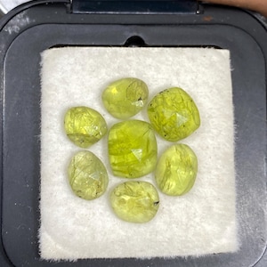 Puede incluir: Un conjunto de seis piedras preciosas de color verde lima, facetadas y sueltas, dispuestas en un patrón circular sobre una almohadilla de fieltro blanco dentro de un estuche de plástico negro. Cada piedra es translúcida con inclusiones.