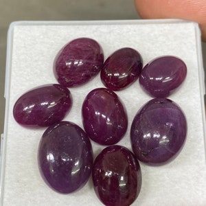 Może przedstawiać: Kolekcja ośmiu owalnych, wypolerowanych kamieni szlachetnych w odcieniach głębokiej czerwieni i fioletu. Kamienie są prezentowane w białym, prostokątnym pojemniku, ukazując ich gładkie, odbijające światło powierzchnie. Prawdopodobnie rubiny, idealne do biżuterii.