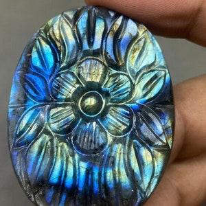 May include: Oval-shaped labradorite gemstone carved with a floral design. The stone features a play of color with blues, greens, and yellows. The carved details create a textured surface, enhancing the gemstone's natural beauty. The gemstone is held in a hand.