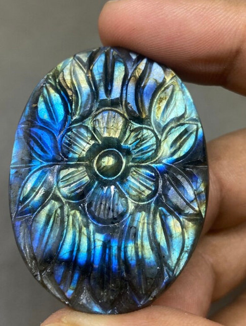 May include: Oval-shaped labradorite gemstone carved with a floral design. The stone features a play of color with blues, greens, and yellows. The carved details create a textured surface, enhancing the gemstone's natural beauty. The gemstone is held in a hand.