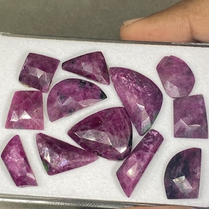 Può includere: Una collezione di undici gemme sfaccettate dai toni magenta profondo e viola con forme geometriche irregolari, inclusi triangoli, trapezi e semicerchi, disposte su un vassoio espositore bianco con visibili strutture cristalline.